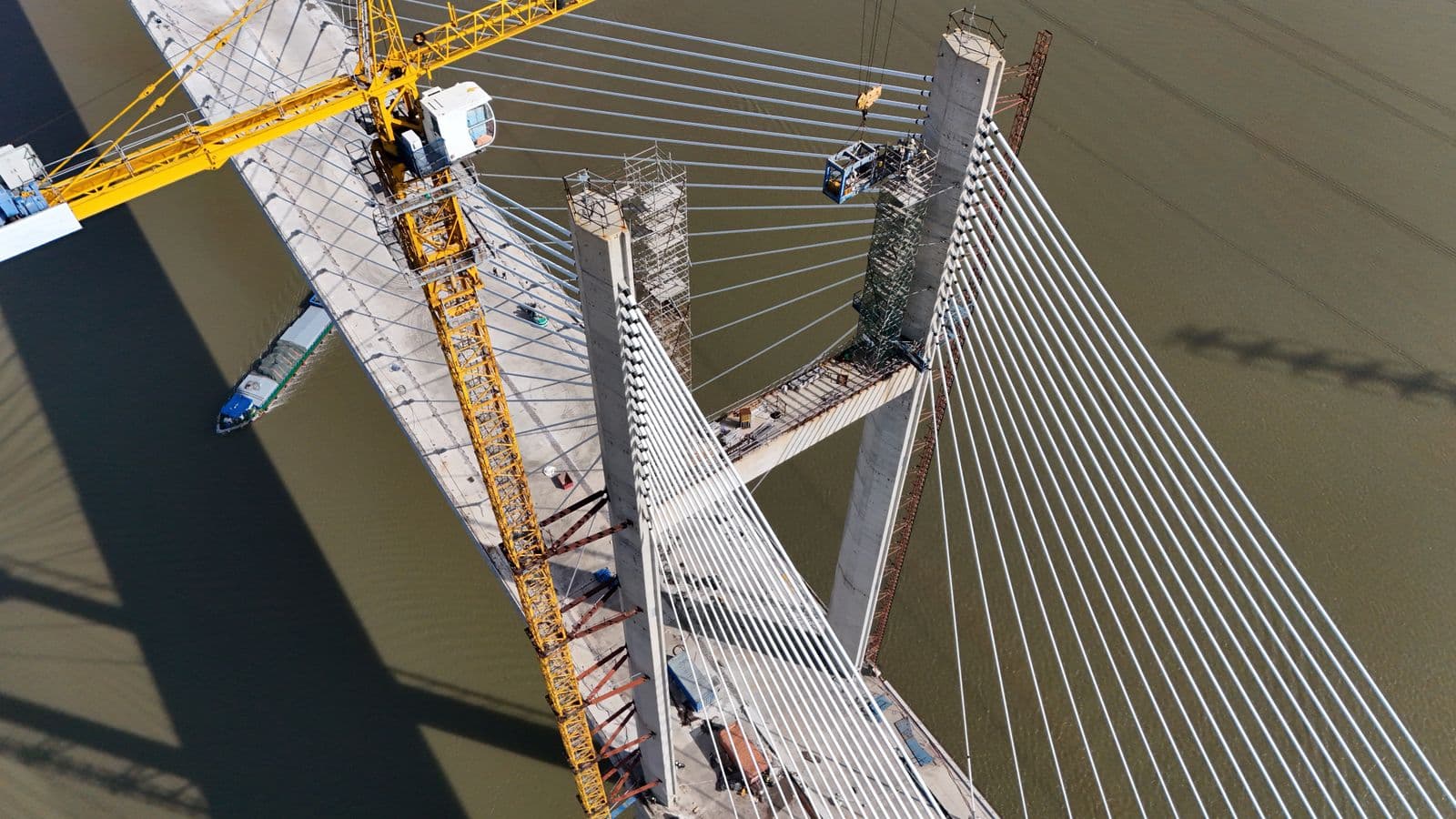 Vista aérea de uma grande ponte estaiada em construção sobre um rio, com guindastes trabalhando nas torres