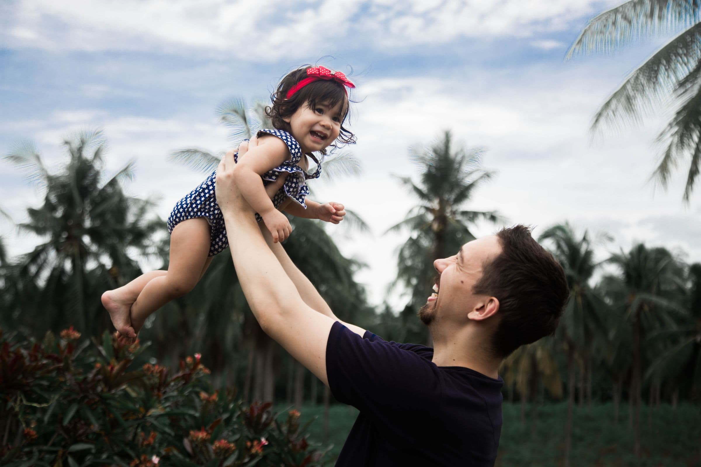 Pai erguendo a filha no colo, os dois sorrindo, em um jardim de coqueiros — um momento comum que vale a pena proteger.
